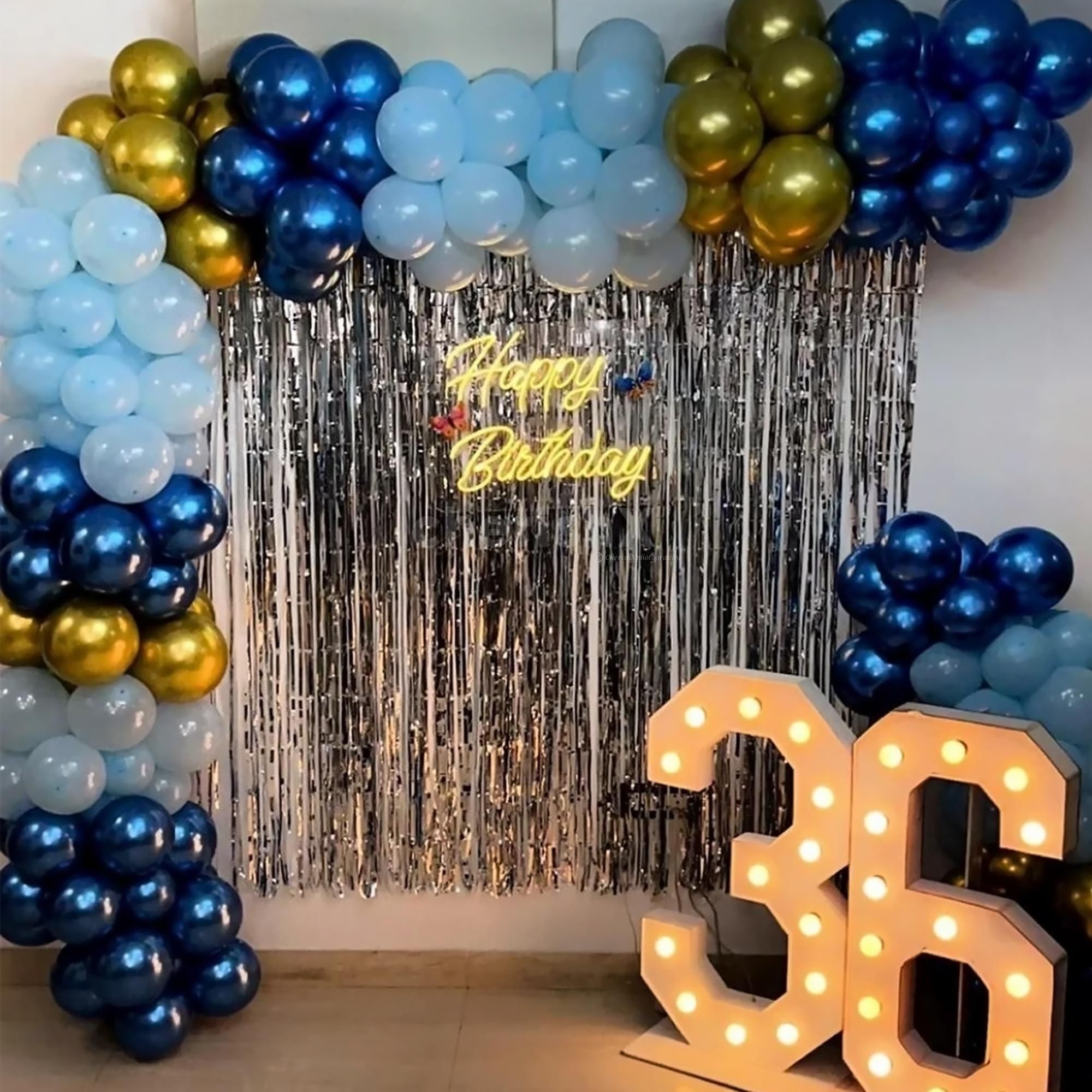 A striking Birthday Decoration featuring a large organic balloon arch in dark blue, light blue, and metallic gold, set against a silver tinsel curtain, with a neon "Happy Birthday" sign and illuminated marquee numbers "36."