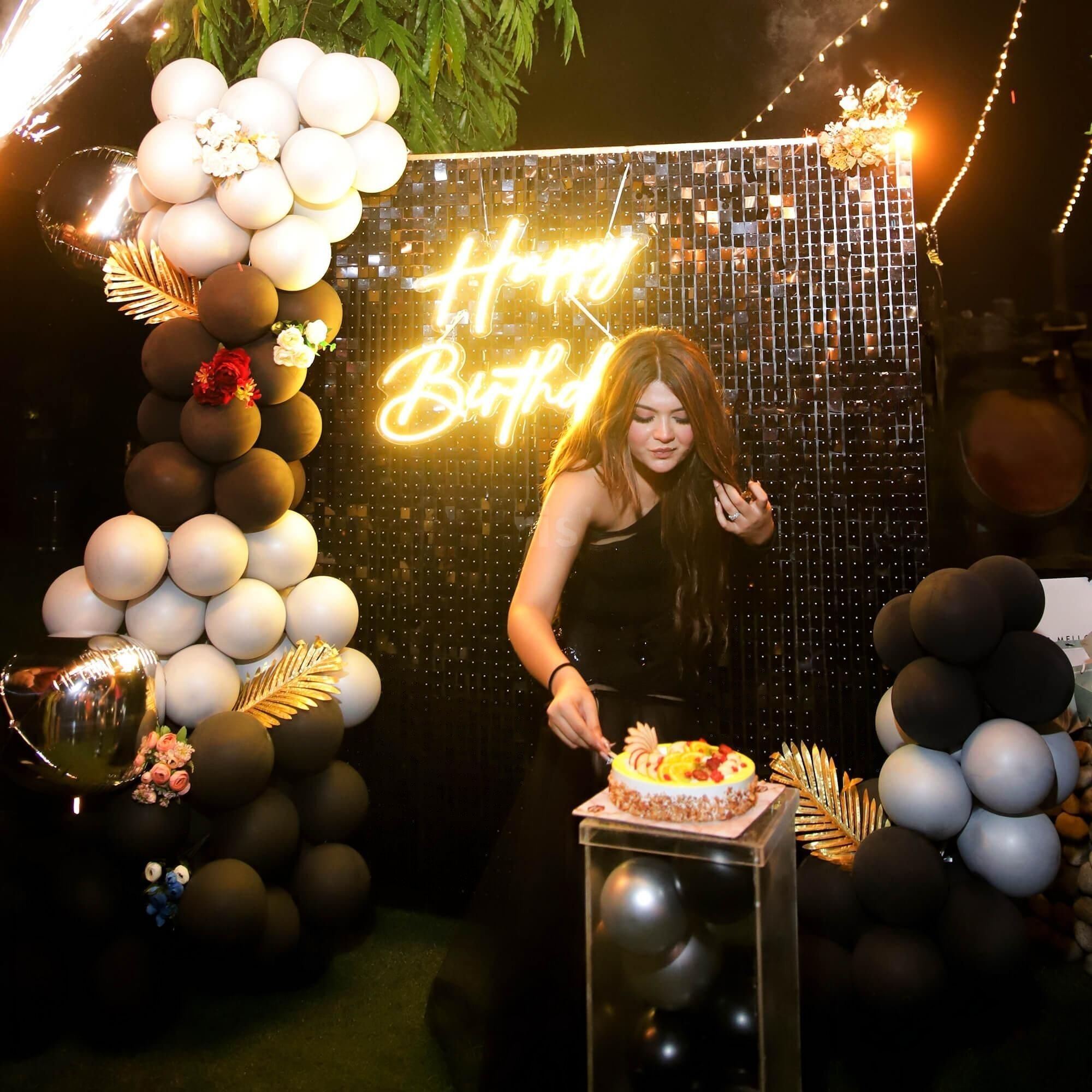 A young woman in a black dress cuts a fruit-topped cake during a nighttime birthday party. The background features a shimmering black sequin wall with a neon "Happy Birthday" sign in orange-yellow cursive, flanked by large, organic balloon arches in black, white, and bronze/brown.