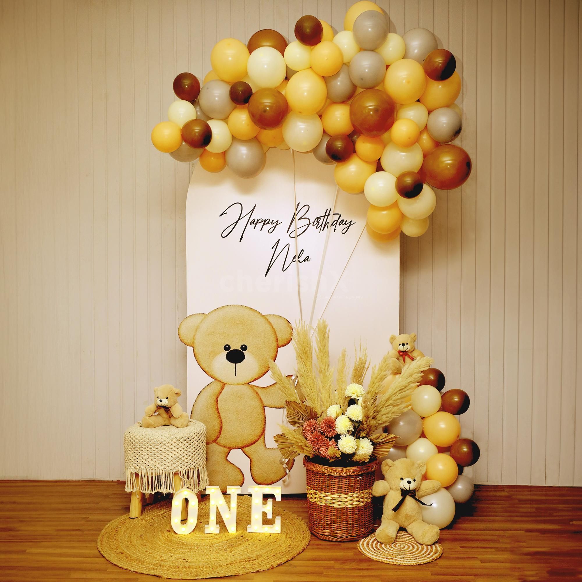 An elegant indoor birthday decoration with an arch-shaped backdrop featuring the word "BIRTHDAY". The setup includes a large organic balloon garland in shades of brown, beige, and white, a oversized teddy bear, and three white cylindrical pedestals, all placed on a patterned carpet against a textured wall with framed artwork and a chandelier.