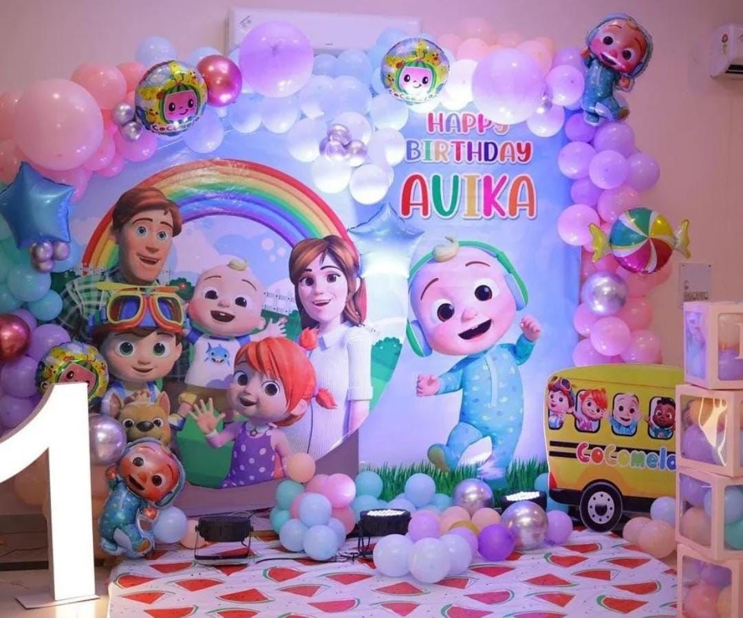 A vibrant Cocomelon-themed first birthday party decoration in Gurgaon, featuring a large backdrop of the Cocomelon family under a rainbow with "HAPPY BIRTHDAY AVIKA". The scene is framed by a pastel balloon arch, with an illuminated number '1' and a Cocomelon school bus cutout.
