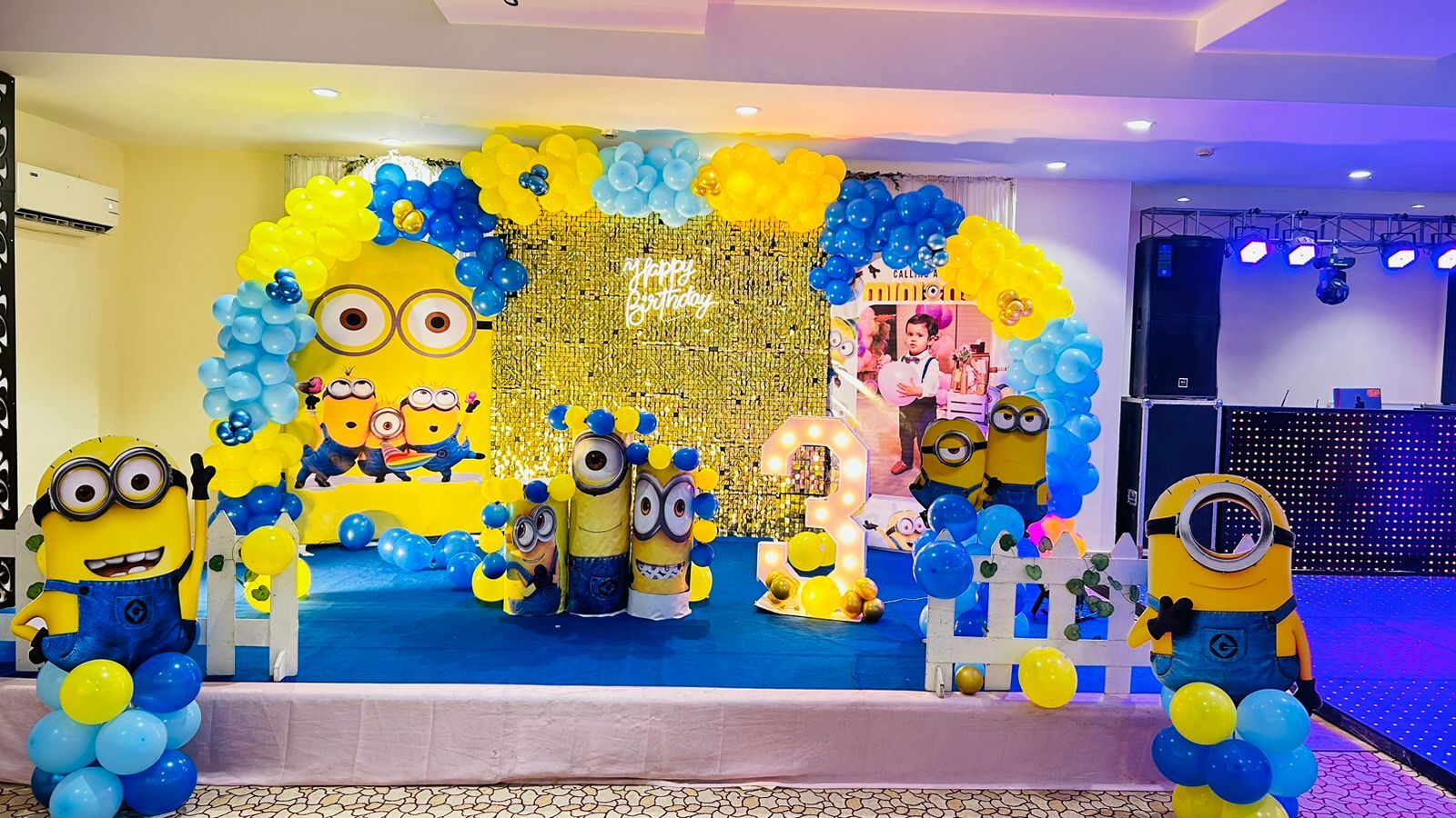 Detailed Minion Theme Birthday Decoration in Gurgaon with a custom character backdrop, yellow and blue balloon clouds, and multiple themed cakes by Adore Celebration.