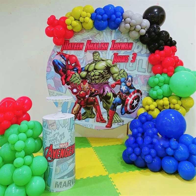A vibrant Avengers Kids Birthday Decor setup featuring a large circular backdrop with Hulk, Iron Man, Spider-Man, and Captain America, framed by a balloon garland in red, blue, green, yellow, and black, for a child turning 3.