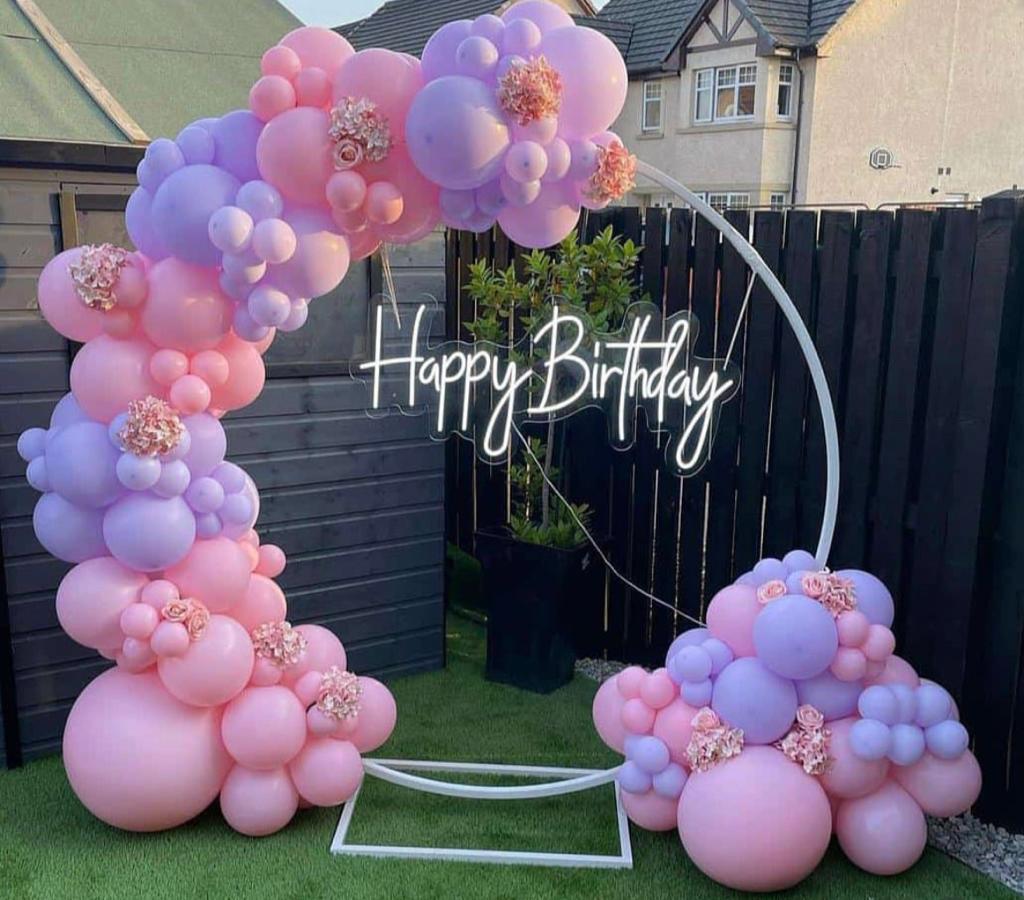 A whimsical Birthday Balloon Ring Setup outdoors, featuring an asymmetrical balloon garland in pastel pink and lavender, accented with small flowers, a white round frame, and a neon "Happy Birthday" sign.