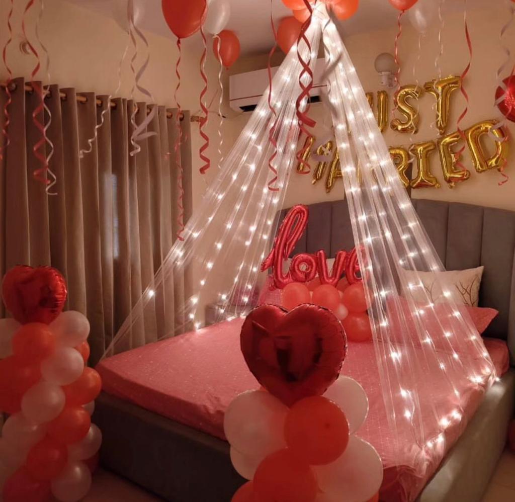 A cozy bedroom decorated for a romantic celebration with a theme of red, white, and orange. The bed features a shimmering sheer canopy draped with fairy string lights, creating an illuminated, intimate tent effect. Above the bed, gold foil balloon letters spell out "JUST MARRIED," and a cursive foil balloon reads "love." Red and white balloons are clustered on the floor in columns and scattered from the ceiling.